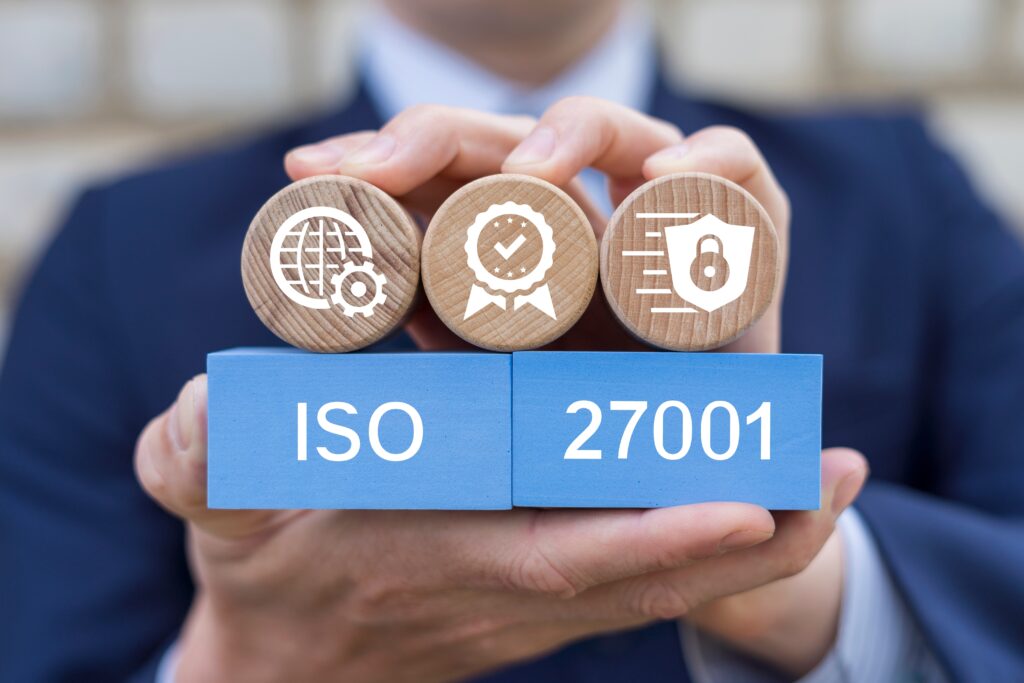 Hände halten blaue Bausteine mit der Aufschrift ISO 27001, ergänzt durch Symbole für Sicherheit, Zertifizierung und Datenschutz