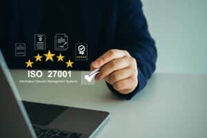 Person setzt ein Häkchen bei ISO 27001, umgeben von Symbolen für Informationssicherheitsmanagement, Audit und Zertifizierung.